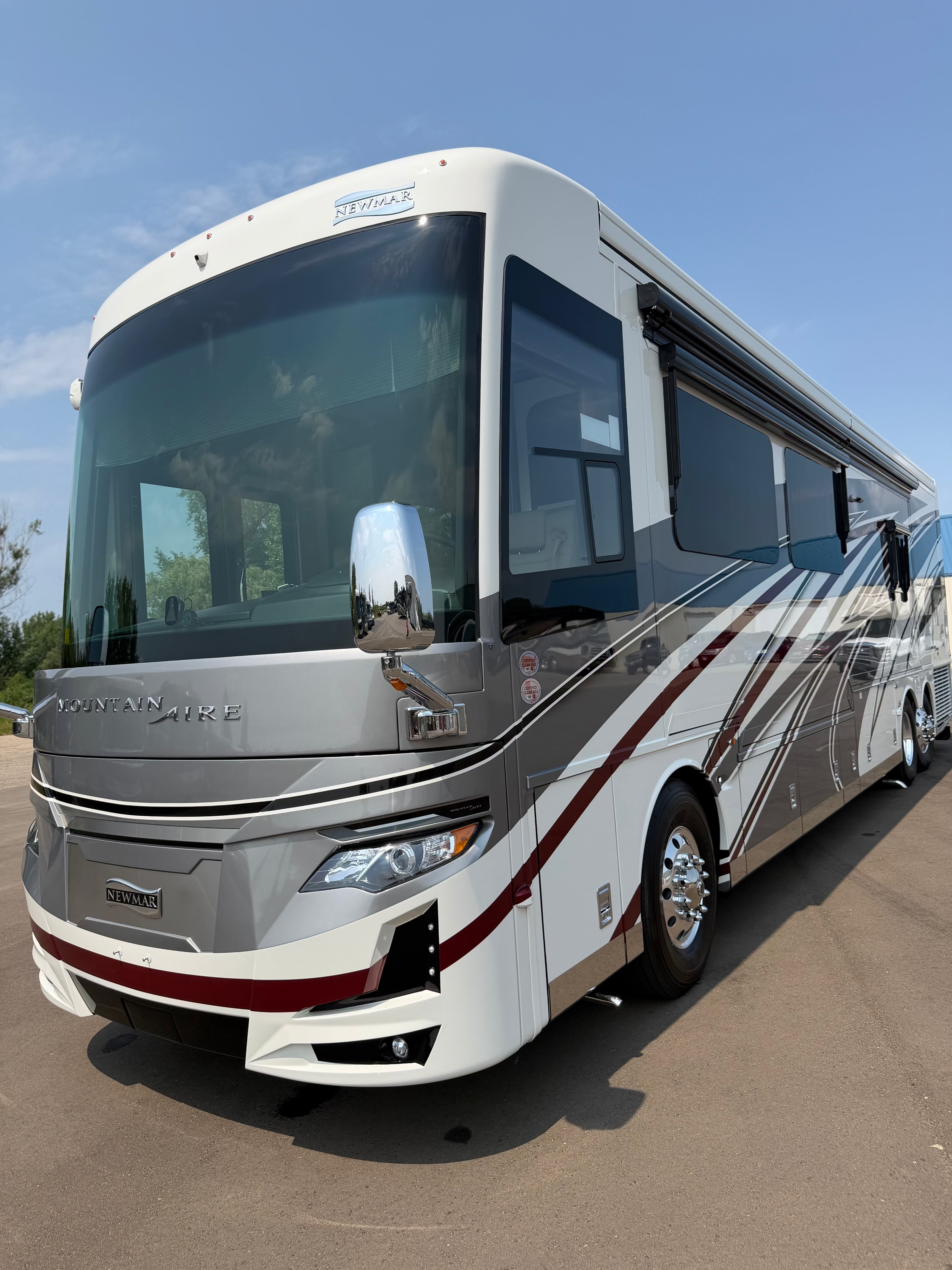 A white and silver motorhome RV with burgundy graphics, photographed outdoors on a paved surface under a blue sky. The front grille shows the RV brand name and model styling.