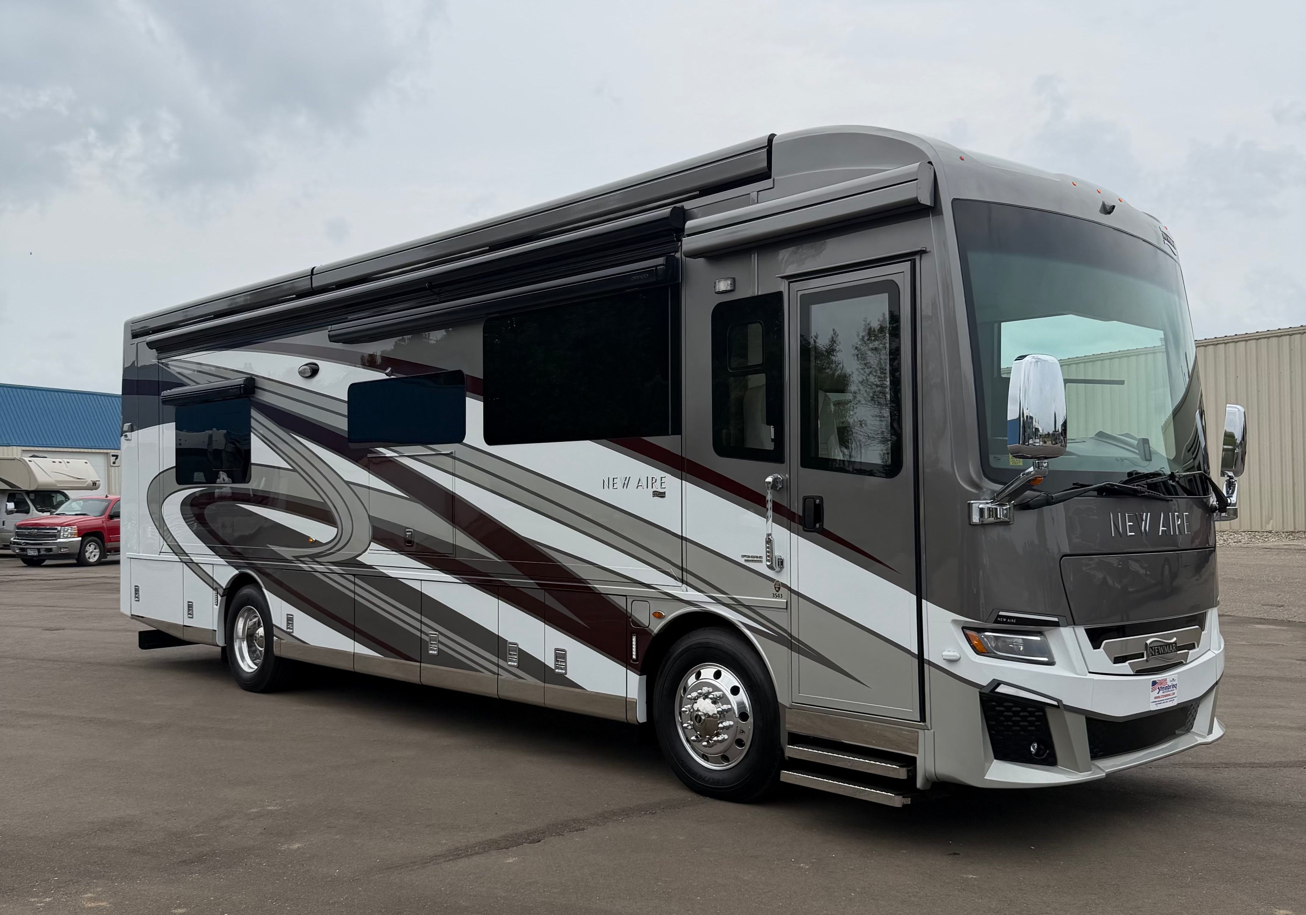 A luxury motorhome RV with a dark gray and silver body and brown graphic stripes, parked outdoors on a paved lot under an overcast sky.