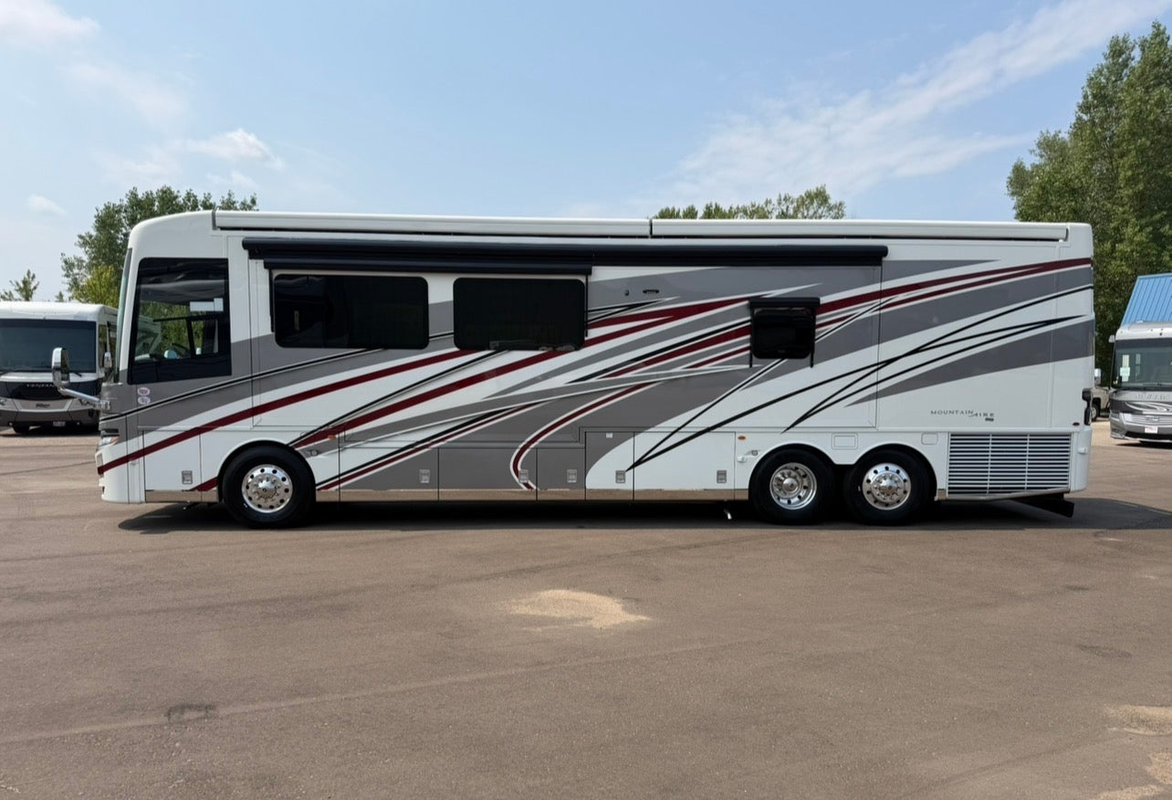 A large motorhome RV with a white body and gray, black, and red graphic stripes, parked outdoors on a paved lot under a blue sky.